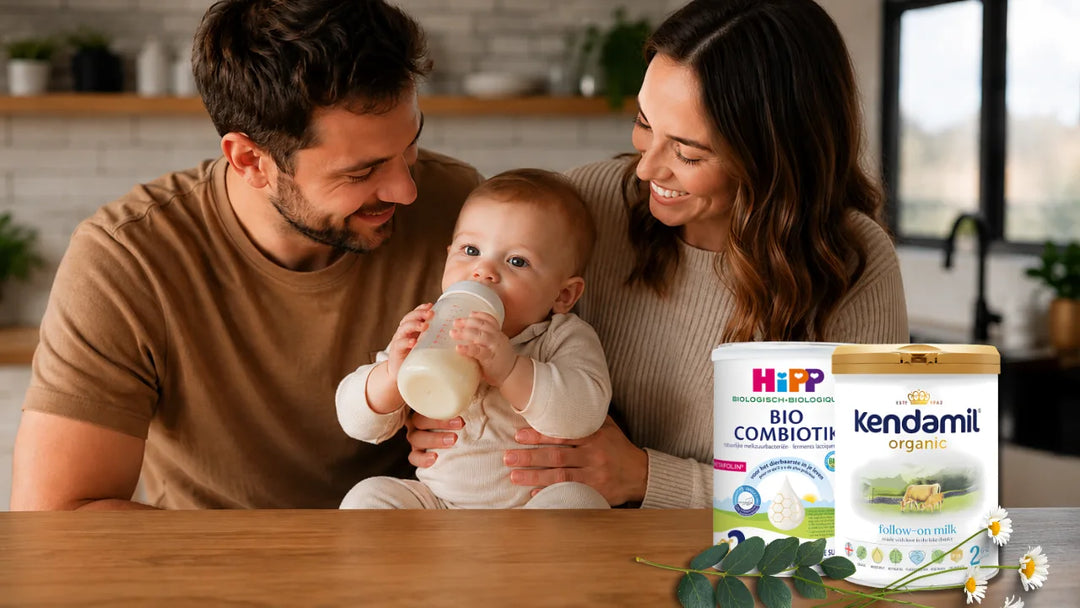 Happy family feeding their baby with a bottle beside HiPP Dutch Stage 2 and Kendamil Organic Stage 2 European baby formula cans in a bright kitchen.